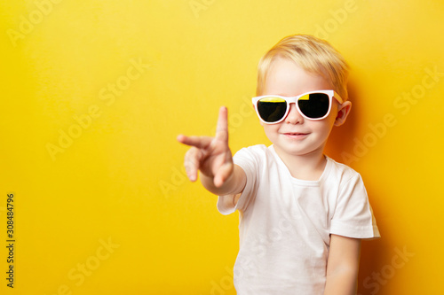 Portrait of cheerful little boy in a white t-shirt wearing sunglasses and looking away on orange background showing two fingers peace sign