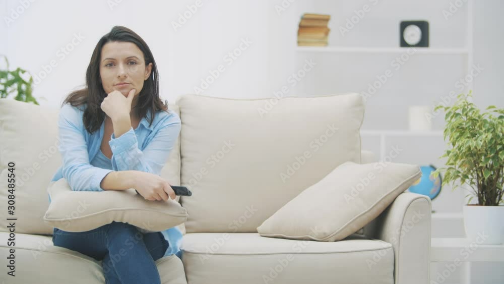 Slowmo. Attractive woman is bored by watching TV, sitting on sofa at home. Suddenly she hears surprising information and smiling contentedly. Copy space. 4K. Slowmo. Attractive woman is bored by watching TV, sitting on sofa at home. Suddenly she hears surprising information and smiling contentedly. Copy space. 4K.