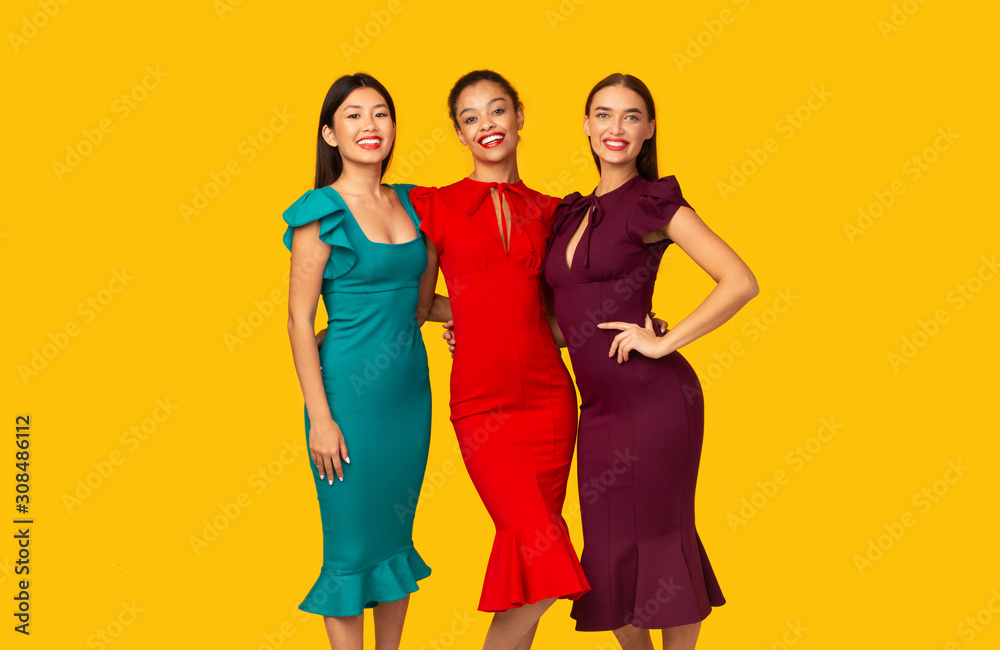 Three Multiracial Models In Dresses Standing Over Yellow Studio Background Three Multiracial Models In Dresses Standing Over Yellow Studio Background