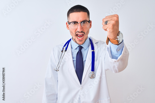 Young doctor man wearing stethoscope over isolated background angry and mad raising fist frustrated and furious while shouting with anger. Rage and aggressive concept.