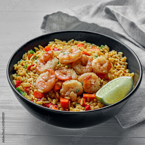 Delicious fried rice with shrimps, lime and vegetables in a black bowl. Traditional Asian food on a white wooden table with a fabric tablecloth.