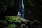 Young Caucasian woman meditating, practicing yoga at waterfall. Gyan mudra. Leke Leke waterfall, Bali, Indonesia.