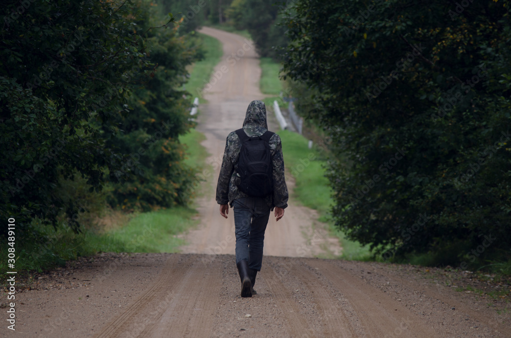 A young man with a backpack travels alone on a dirt road in cloudy gloomy weather A young man with a backpack travels alone on a dirt road in cloudy gloomy weather