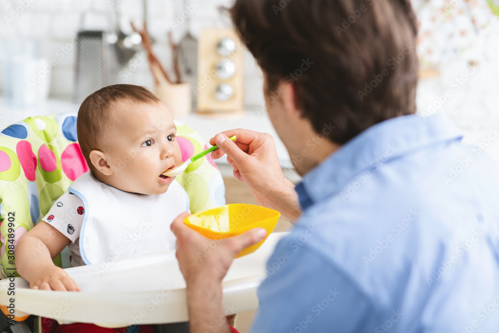 Father feeding his curious baby son at kitchen interior Father feeding his curious baby son at kitchen interior