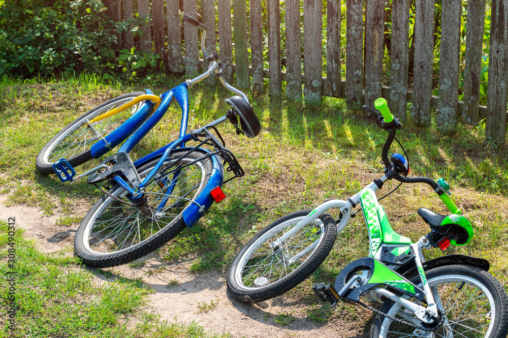 Fototapeta premium Children's and adult bicycles lay in the grass on the side of the road near a rural fence. Country trips on the weekend