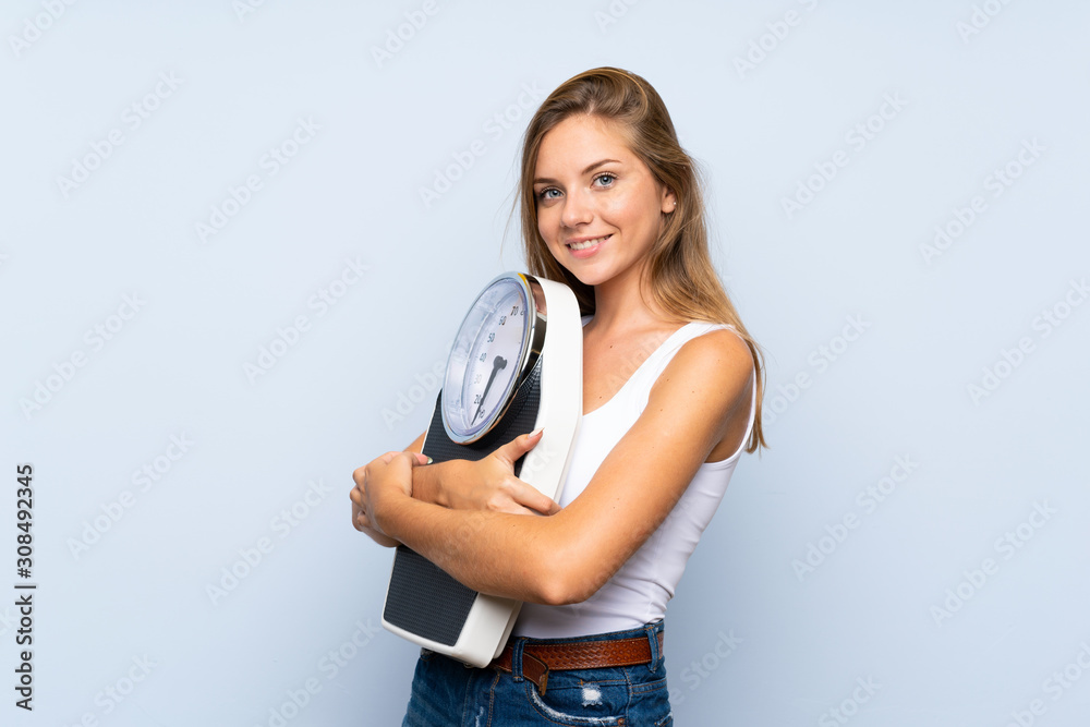 Young blonde girl with weighing machine over isolated blue white background Young blonde girl with weighing machine over isolated blue white background
