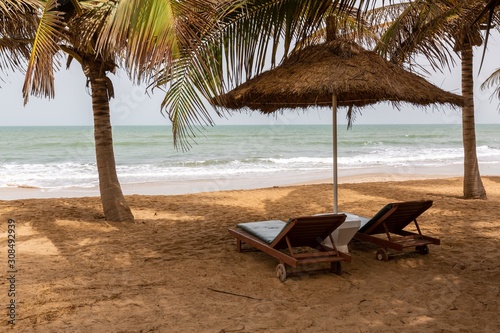 Fototapeta Naklejka Na Ścianę i Meble -  Beach in the Gambia with thatch umbrellas palms and beach chairs with the sea on the background