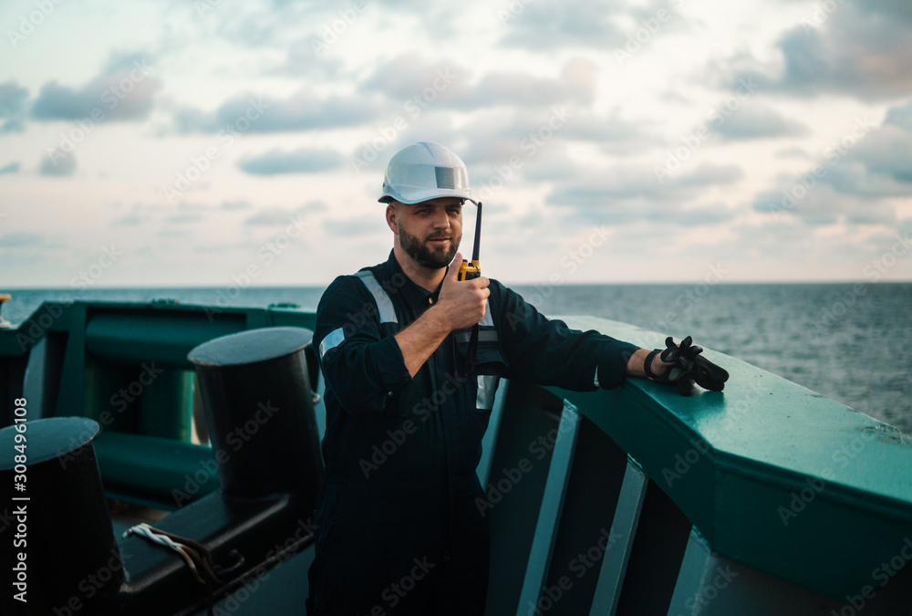 Obraz premium Marine Deck Officer or Chief mate on deck of offshore vessel or ship , wearing PPE personal protective equipment - helmet, coverall. He holds VHF walkie-talkie radio in hands.