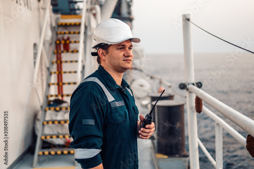 Marine Deck Officer or Chief mate on deck of offshore vessel or ship , wearing PPE personal protective equipment - helmet, coverall. He holds VHF walkie-talkie radio in hands. photo