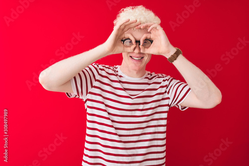 Young albino blond man wearing striped t-shirt and glasses over isolated red background doing ok gesture like binoculars sticking tongue out, eyes looking through fingers. Crazy expression.