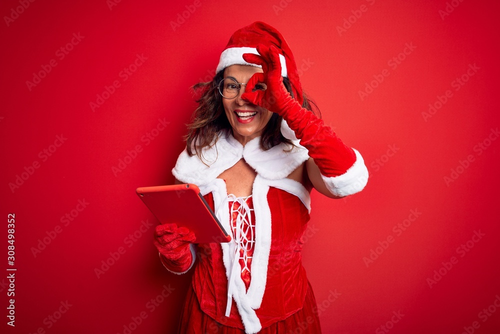 Middle age woman wearing Santa Claus costume using tablet over isolated red background with happy face smiling doing ok sign with hand on eye looking through fingers Middle age woman wearing Santa Claus costume using tablet over isolated red background with happy face smiling doing ok sign with hand on eye looking through fingers