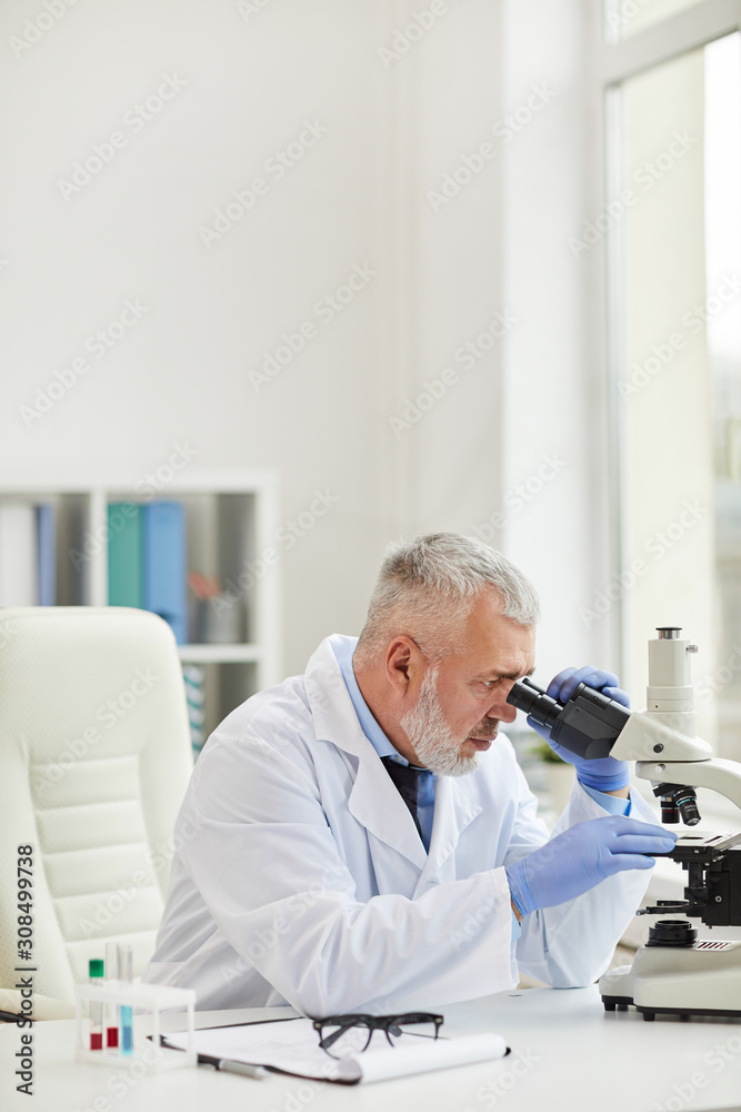 Mature doctor with grey hair sitting at his workplace and using microscope in his work while working at office Mature doctor with grey hair sitting at his workplace and using microscope in his work while working at office
