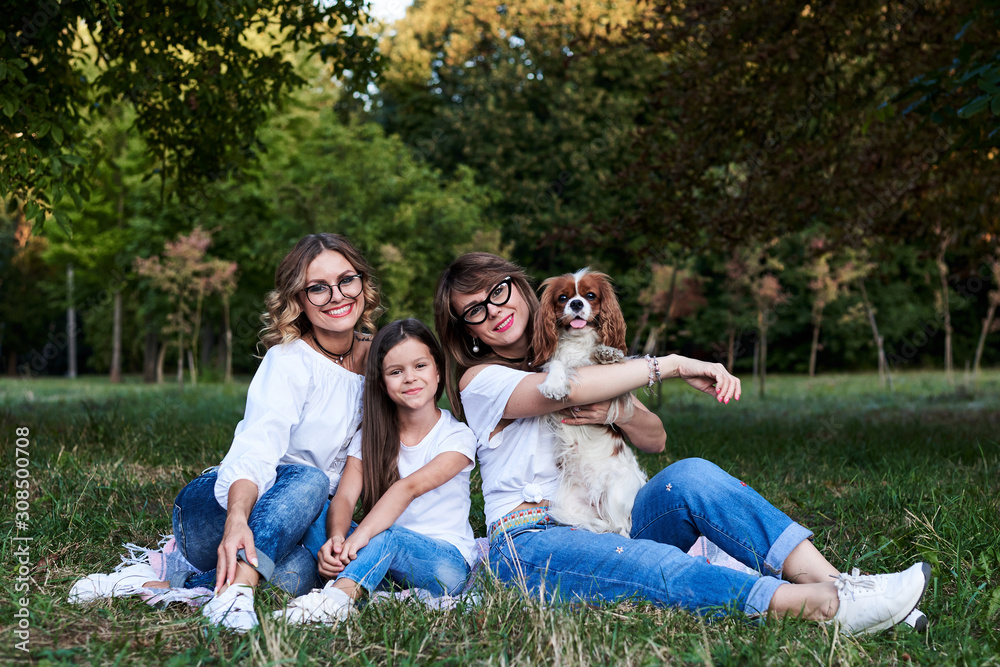 Two young blond women and one brunette girl, wearing jeans and white t-shirts, sitting on green grass in park, playing with cavalier king charles spaniel, smiling, laughing. Leisure time in summer. Two young blond women and one brunette girl, wearing jeans and white t-shirts, sitting on green grass in park, playing with cavalier king charles spaniel, smiling, laughing. Leisure time in summer.