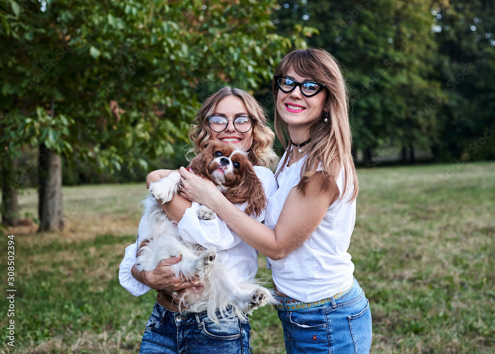 Two young blond women, wearing white t-shirts,, holding small white and brown dog in park in summer, smiling. Cavalier king charles spaniel with his owners on a walk.