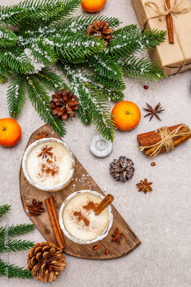 Eggnog with cinnamon and nutmeg for Christmas and winter holidays. Homemade beverage in glasses with spicy rim. Tangerines, candles, gift. Stone concrete background