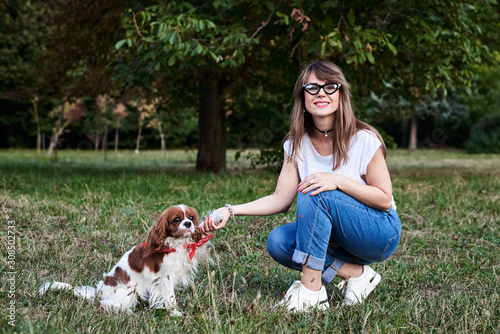 Young blond woman, kneeling down, holding her dog's paw in park in summer. Dog owner training her Cavalier king charles spaniel.