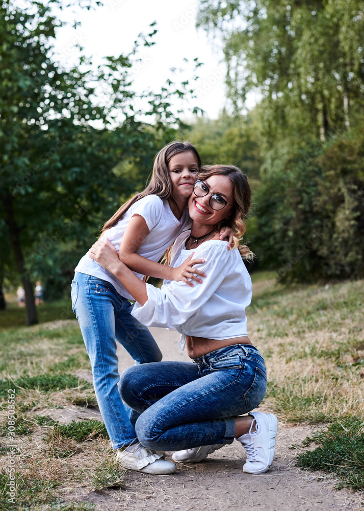 Young blond mother hugging her brunette daughter in the park in summer. Woman and girl, wearing blue jeans and white t-shirts, embracing, looking at each other, smiling. Family leisure fun time. Young blond mother hugging her brunette daughter in the park in summer. Woman and girl, wearing blue jeans and white t-shirts, embracing, looking at each other, smiling. Family leisure fun time.