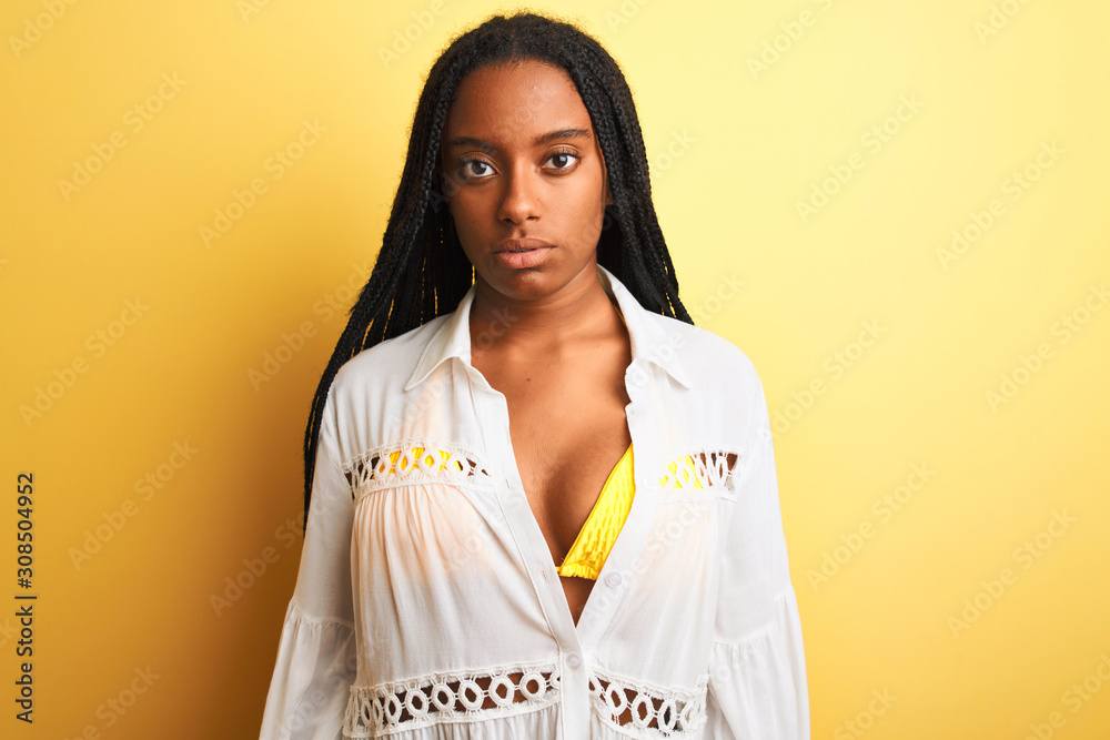 Fototapeta premium African american woman on vacation wearing bikini and shirt over isolated yellow background with serious expression on face. Simple and natural looking at the camera.