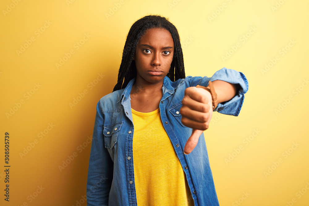 Young african american woman wearing denim shirt standing over isolated yellow background looking unhappy and angry showing rejection and negative with thumbs down gesture. Bad expression. Young african american woman wearing denim shirt standing over isolated yellow background looking unhappy and angry showing rejection and negative with thumbs down gesture. Bad expression.