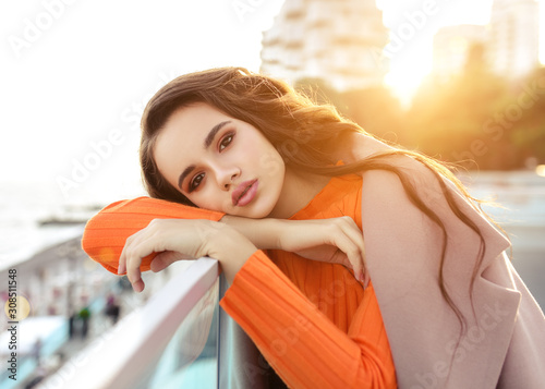 Beautiful young girl with dark hair stands near the sea in a beige coat and smiles. Sunset on the seefront. photo