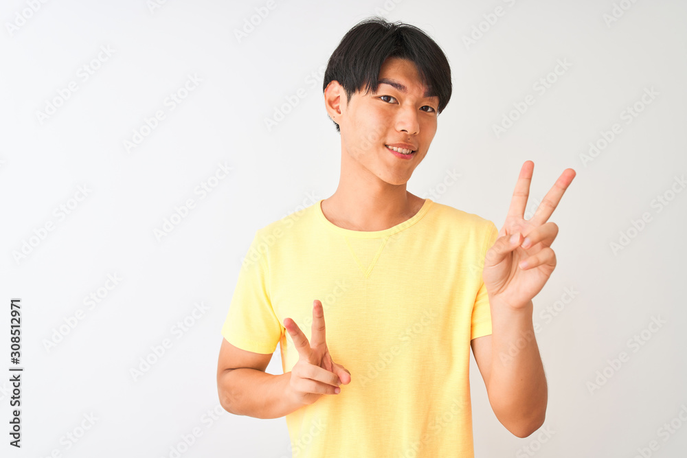 Chinese man wearing yellow casual t-shirt standing over isolated white background smiling looking to the camera showing fingers doing victory sign. Number two. Chinese man wearing yellow casual t-shirt standing over isolated white background smiling looking to the camera showing fingers doing victory sign. Number two.