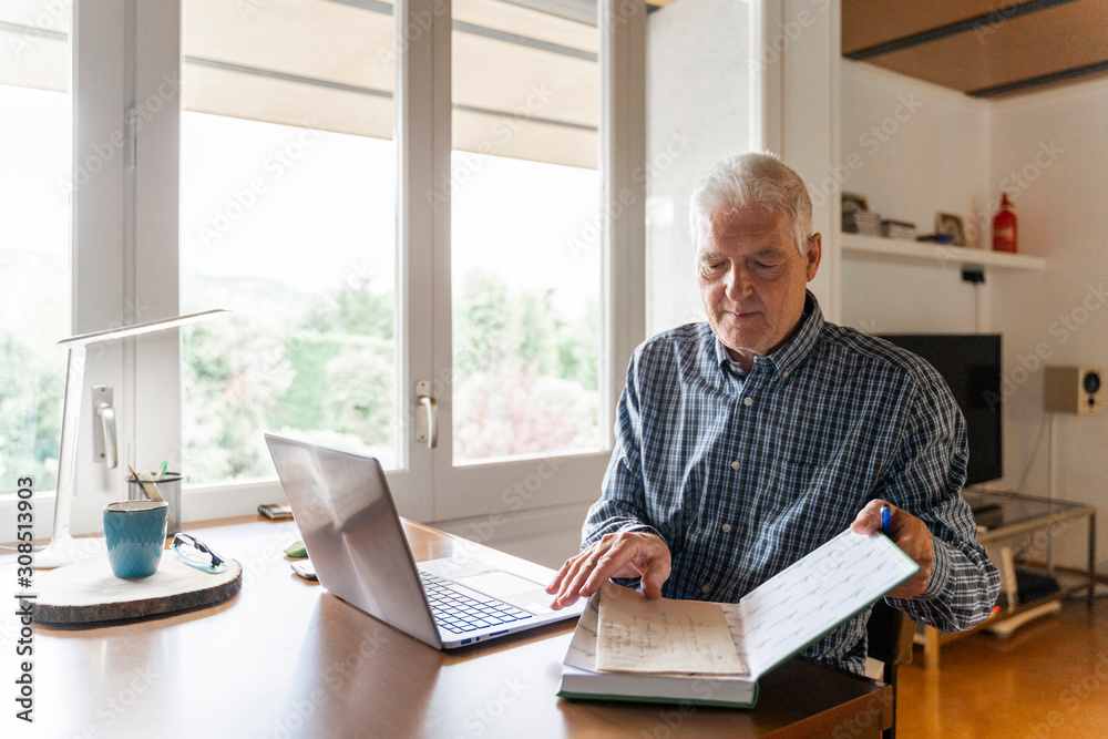 Senior man sitting at home in front of laptop turning pages in notebook Senior man sitting at home in front of laptop turning pages in notebook
