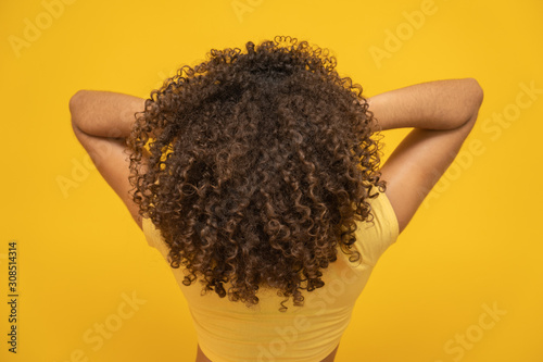 Backwards American African woman with her curly hair on yellow background. Laughing curly woman in sweater touching her hair and looking at the camera.
