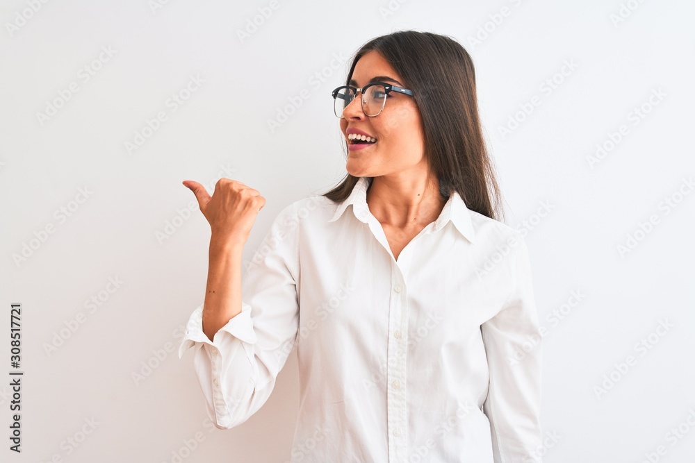 Young beautiful businesswoman wearing glasses standing over isolated white background smiling with happy face looking and pointing to the side with thumb up.