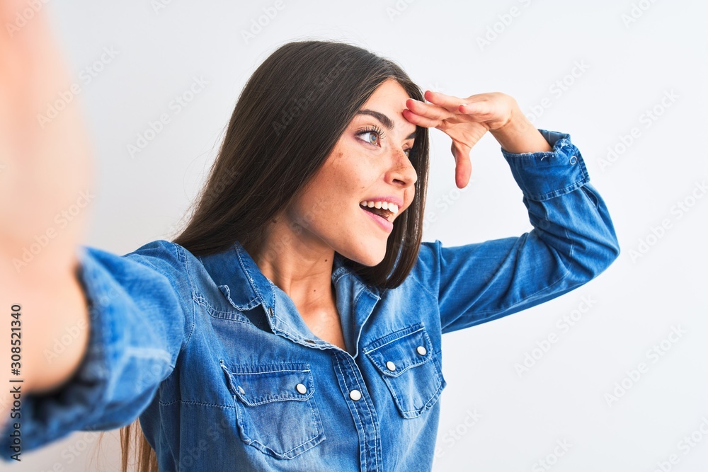 Beautiful woman wearing denim shirt make selfie by camera over isolated white background very happy and smiling looking far away with hand over head. Searching concept. Beautiful woman wearing denim shirt make selfie by camera over isolated white background very happy and smiling looking far away with hand over head. Searching concept.