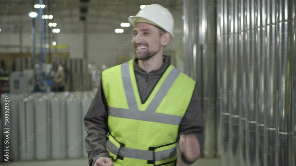 Portrait of cheerful Caucasian male worker in protective helmet dancing at the background of steel metal pipes. Happy worker at his workplace on chimneys production site. Manufacture, steel industry. Portrait of cheerful Caucasian male worker in protective helmet dancing at the background of steel metal pipes. Happy worker at his workplace on chimneys production site. Manufacture, steel industry.