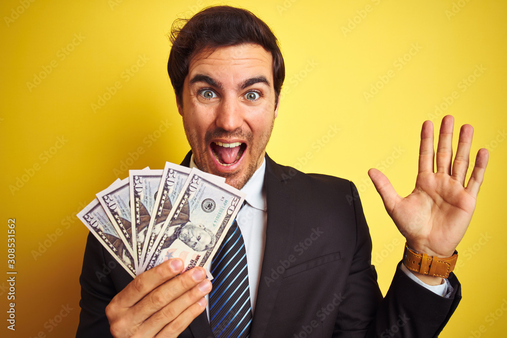 Young handsome businessman wearing suit holding dollars over isolated yellow background very happy and excited, winner expression celebrating victory screaming with big smile and raised hands Young handsome businessman wearing suit holding dollars over isolated yellow background very happy and excited, winner expression celebrating victory screaming with big smile and raised hands
