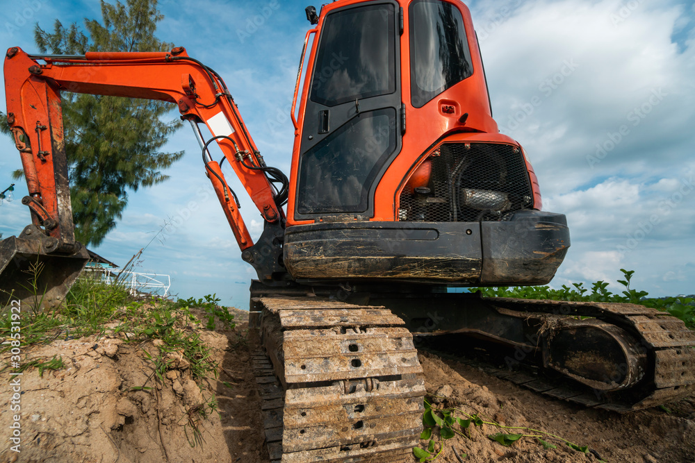 Small orange excavator on a ground against blue sky and sea for a works on construction site. Small tracked excavator standing on a ground with a blue sea on background. Heavy industry. Small orange excavator on a ground against blue sky and sea for a works on construction site. Small tracked excavator standing on a ground with a blue sea on background. Heavy industry.