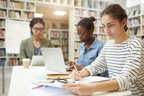 Young woman sitting at the table and working with papers she preparing for exams together with her friends in the background