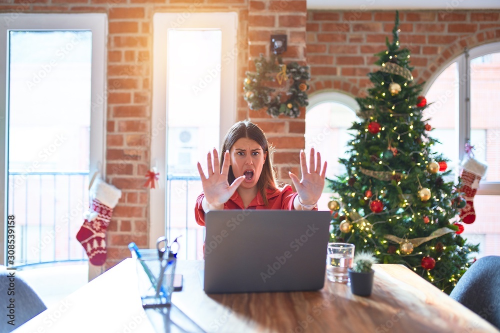 Beautiful woman sitting at the table working with laptop at home around christmas tree doing stop gesture with hands palms, angry and frustration expression Beautiful woman sitting at the table working with laptop at home around christmas tree doing stop gesture with hands palms, angry and frustration expression