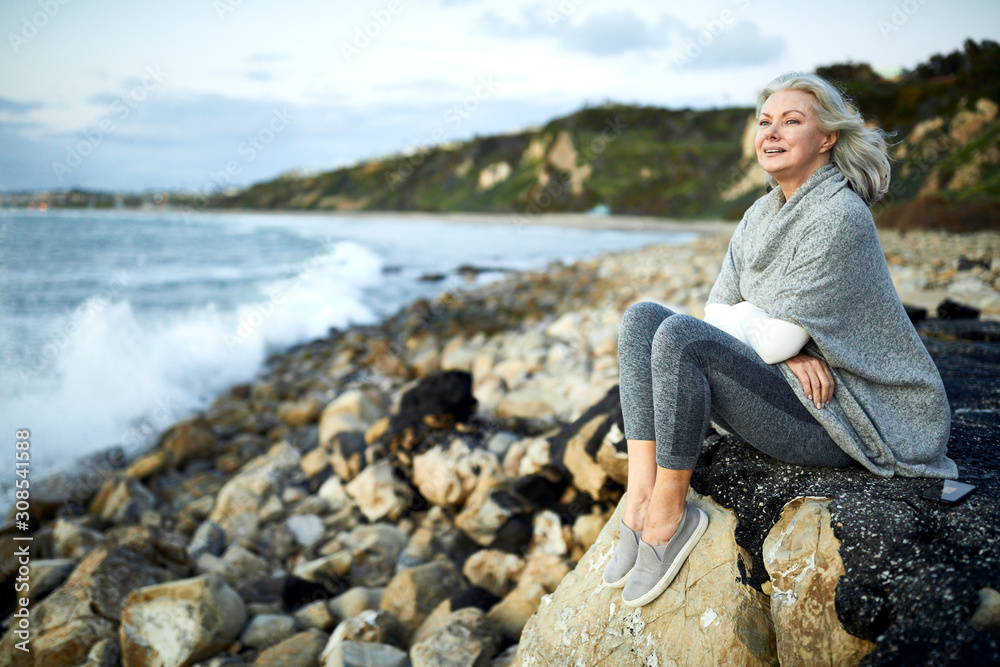 Side view of thoughtful senior woman sitting on rock at Manhattan Beach against sky during sunset Side view of thoughtful senior woman sitting on rock at Manhattan Beach against sky during sunset