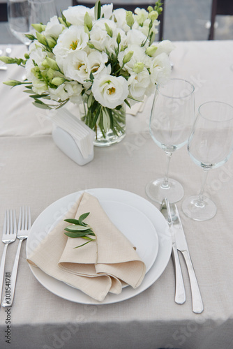 Table setting with beige napkin on empty white plate, wineglasses and cutlery on table, copy space. Place setting at wedding reception. Table served for banquet