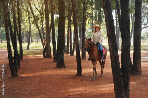Young woman in shirt and straw hat, rides brown horse in the morning park, blurred trees in background