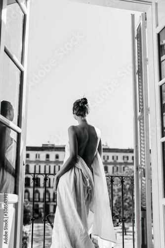 Dziewczyna odpoczywająca w hotelu stoi w otworze otwartego balkonu, owinięta w prześcieradło. Nagie plecy zwrócone do aparatu, zdjęcie czarno-białe