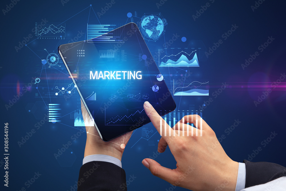 Businessman holding a foldable smartphone with MARKETING inscription, successful business concept Businessman holding a foldable smartphone with MARKETING inscription, successful business concept