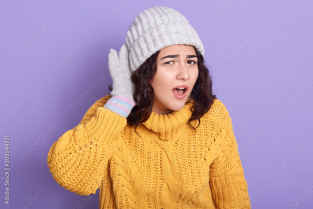Image of curly haired emotional energetic young woman standing isolated over lilac background in studio, opening yellow sweater and hat, gloves, putting hand to ear, trying to hear something. Image of curly haired emotional energetic young woman standing isolated over lilac background in studio, opening yellow sweater and hat, gloves, putting hand to ear, trying to hear something.