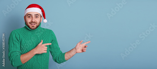 Horizontal studio shot of emotional energetic charismatic man showing direction, making gesture, wearing green sweater and red santa claus hat, being in good mood. Copyspace for advertisement.