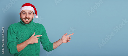 Horizontal studio shot of emotional energetic charismatic man showing direction, making gesture, wearing green sweater and red santa claus hat, being in good mood. Copyspace for advertisement. photo