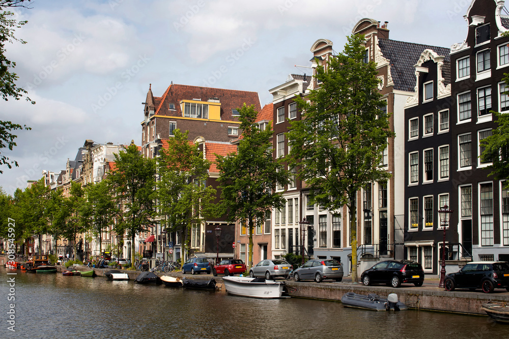 View of canal, parked boats, cars, trees and historical, traditional buildings showing Dutch architecture style in Amsterdam. It is a summer day.