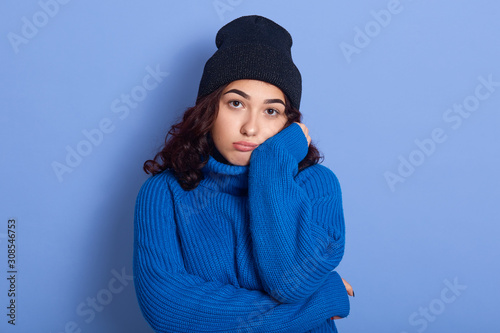 Picture of bored unsatisfied dark haired young female standing isolated over blue background in studio, touching her face with hands, wearing blue hat and sweater. People and boredom concept. photo