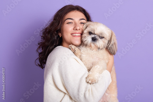 Portrait of positive magnetic young girl holding Maltese in both hands, pressing its close to face, smiling sincerely, looking directly at camera, wearing white sweater. People and pets concept. photo