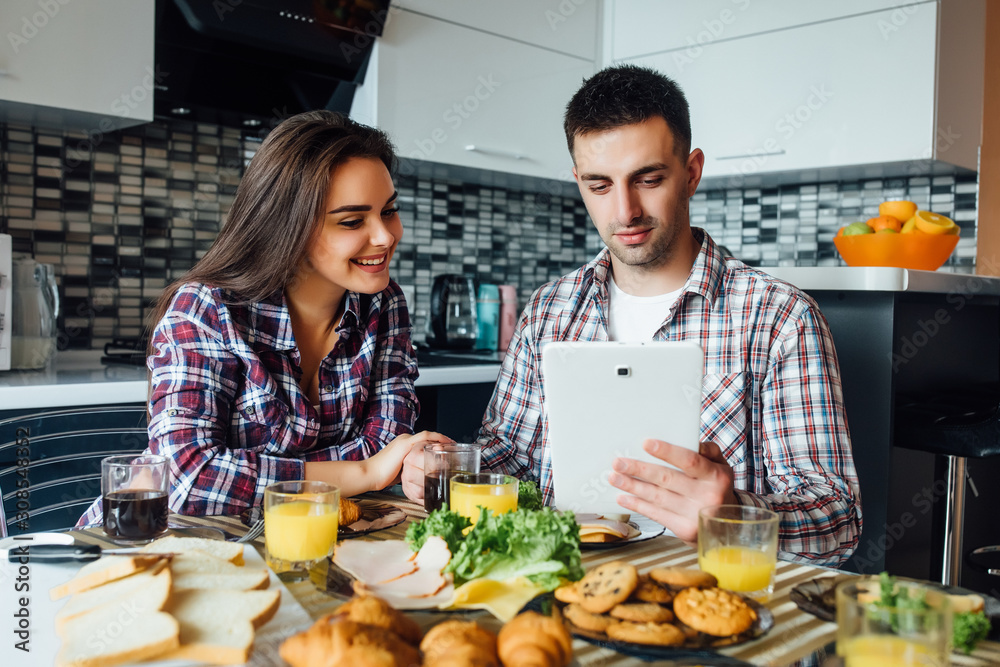 Young attractive couple of man and woman have a breakfast together in morning at kitchen, loooking recipes in internet on tablet computer, smiling.