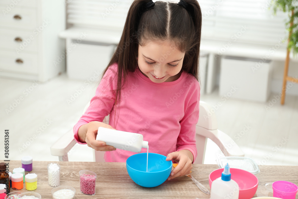 Cute little girl pouring glue into bowl at table in room. DIY slime toy Cute little girl pouring glue into bowl at table in room. DIY slime toy