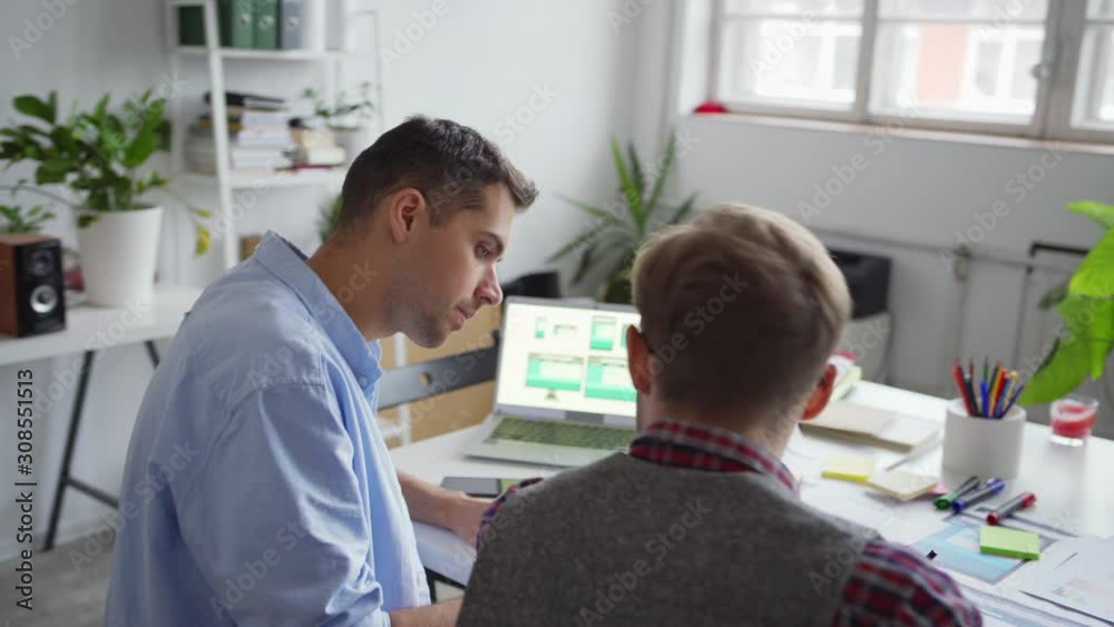 Zoom out panning shot of two web designers working on app interface for various devices. Creative professionals discussing ideas and samples presented on laptop screen sitting at desk in office