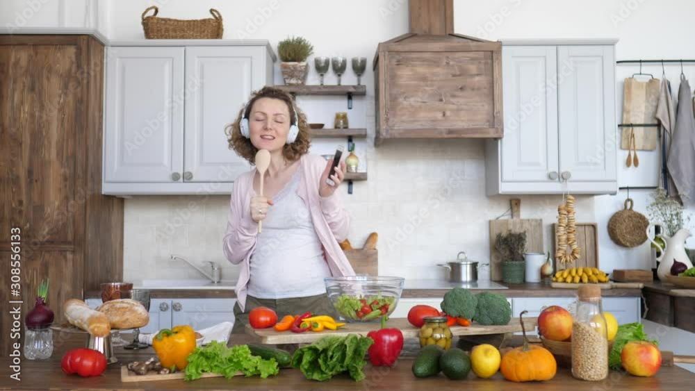 Pregnant Woman Dancing And Singing In Headphones With Mobile Phone On Kitchen. Pregnant Woman Dancing And Singing In Headphones With Mobile Phone On Kitchen.