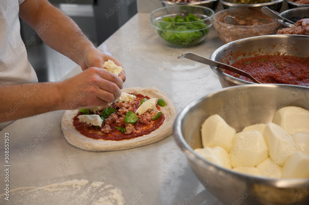 Obraz premium Preparing Pizza Margherita on a marble top. Pizzaiolo puts pieces of sausage, mozzarella and basil leaves over a raw pizza dough, with tomato sauce. Selective focus.